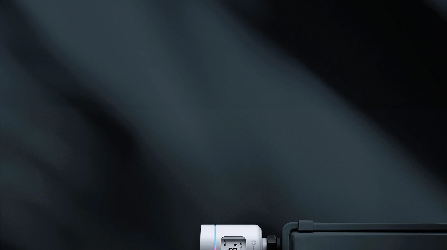 Smart thermostat attached to a black radiator, displaying a temperature of 3.5 degrees. The wall behind is dark with subtle shadows.