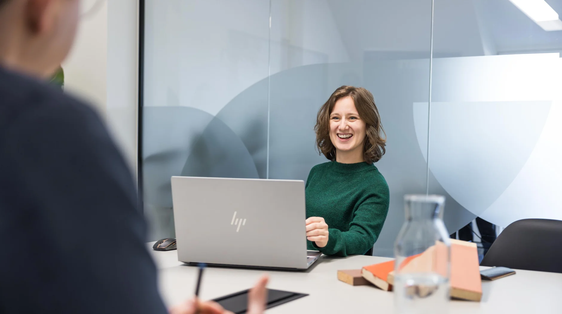 A woman in a green sweater sits at a table with a laptop, smiling at a person opposite her in a modern office setting.