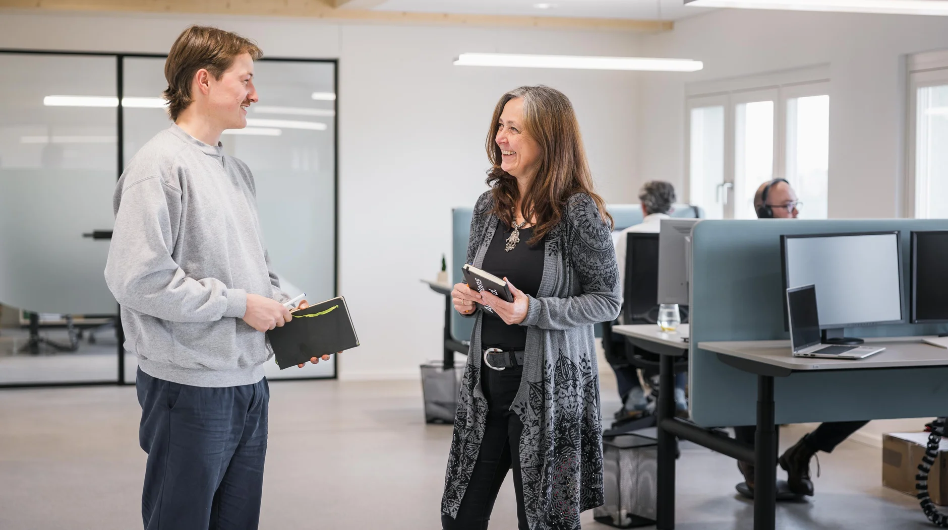 Two people smiling and conversing in a modern office. One holds a notebook, the other a tablet. Desks and computers are visible in the background.