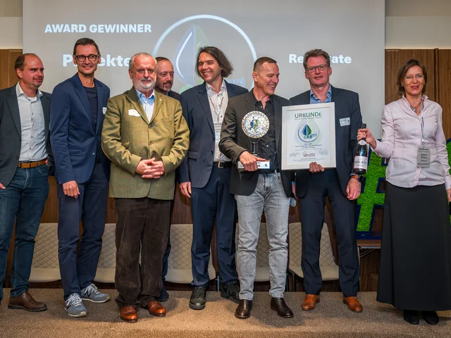 A group of eight people, some holding an award and certificate, stand smiling on a stage with a "WINNER" sign in the background.