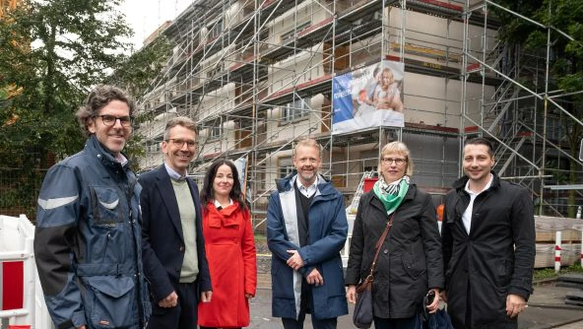 A group of people in coats stand smiling in front of a building under construction with scaffolding.
