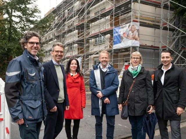 A group of people in coats stand smiling in front of a building under construction with scaffolding.