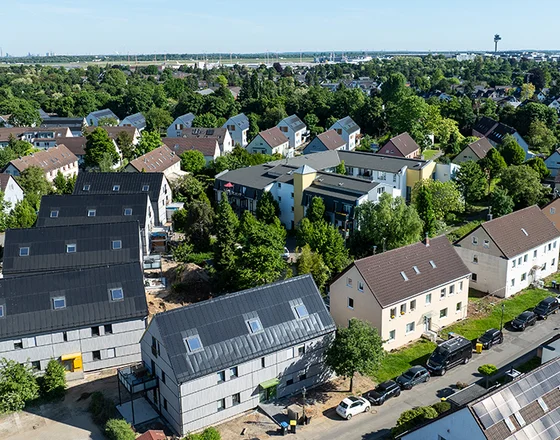 Aerial view of a suburban neighborhood with houses, some under construction, surrounded by trees and greenery on a sunny day.