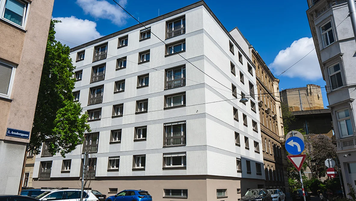 White multi-story building on a sunny day, surrounded by parked cars and trees, with a street sign in the foreground.