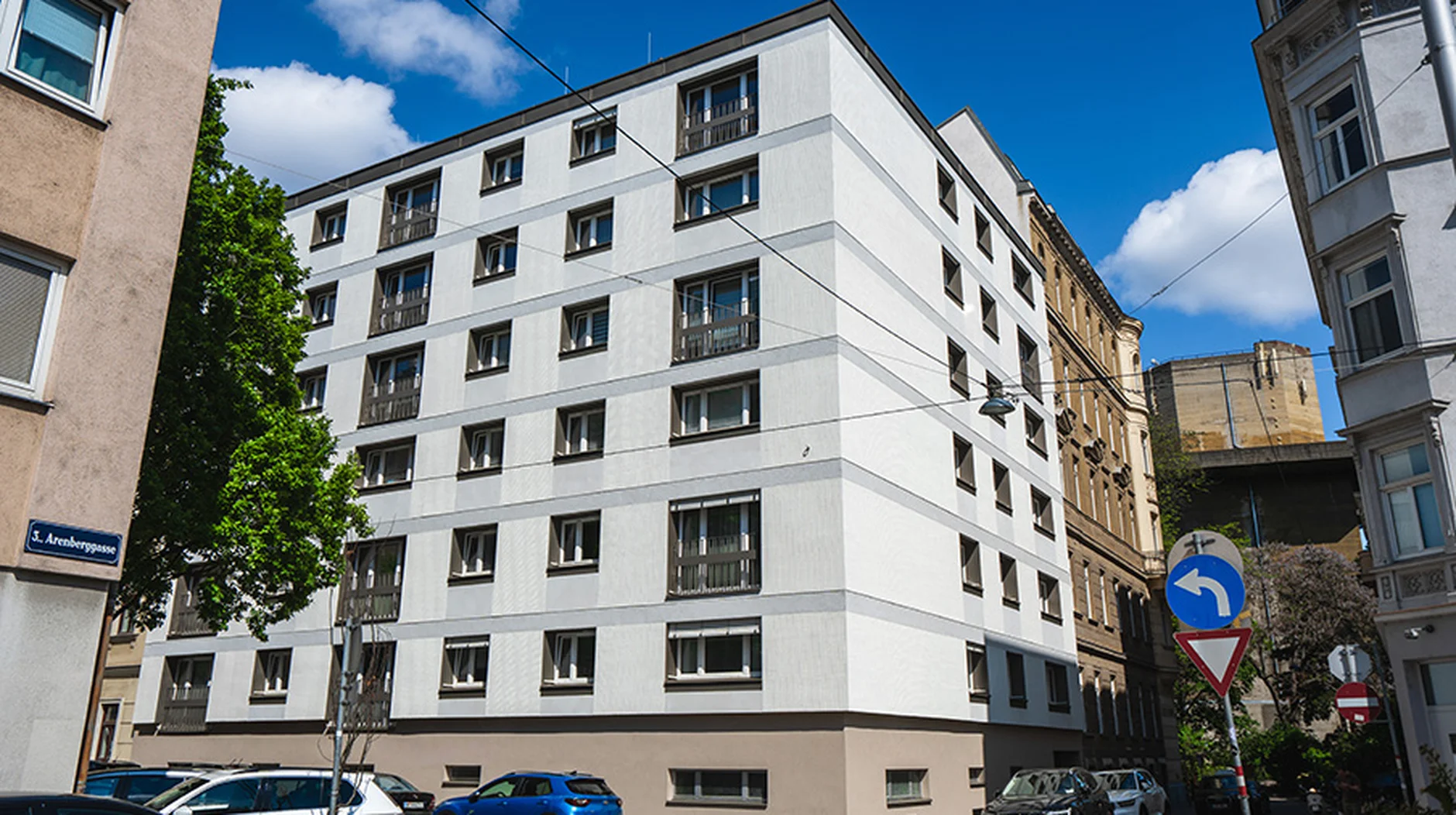 White multi-story building on a sunny day, surrounded by parked cars and trees, with a street sign in the foreground.