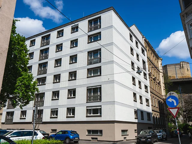 White multi-story building on a sunny day, surrounded by parked cars and trees, with a street sign in the foreground.