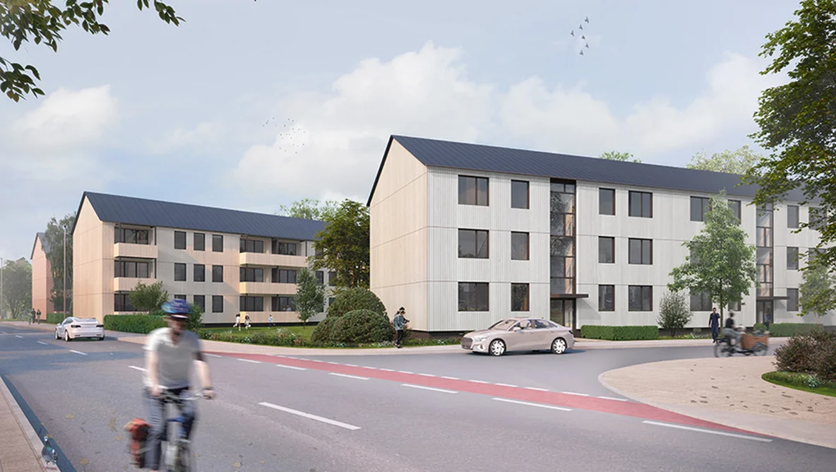 Modern apartment buildings with sloped roofs alongside a road. Cyclist and pedestrians are visible under a partly cloudy sky.