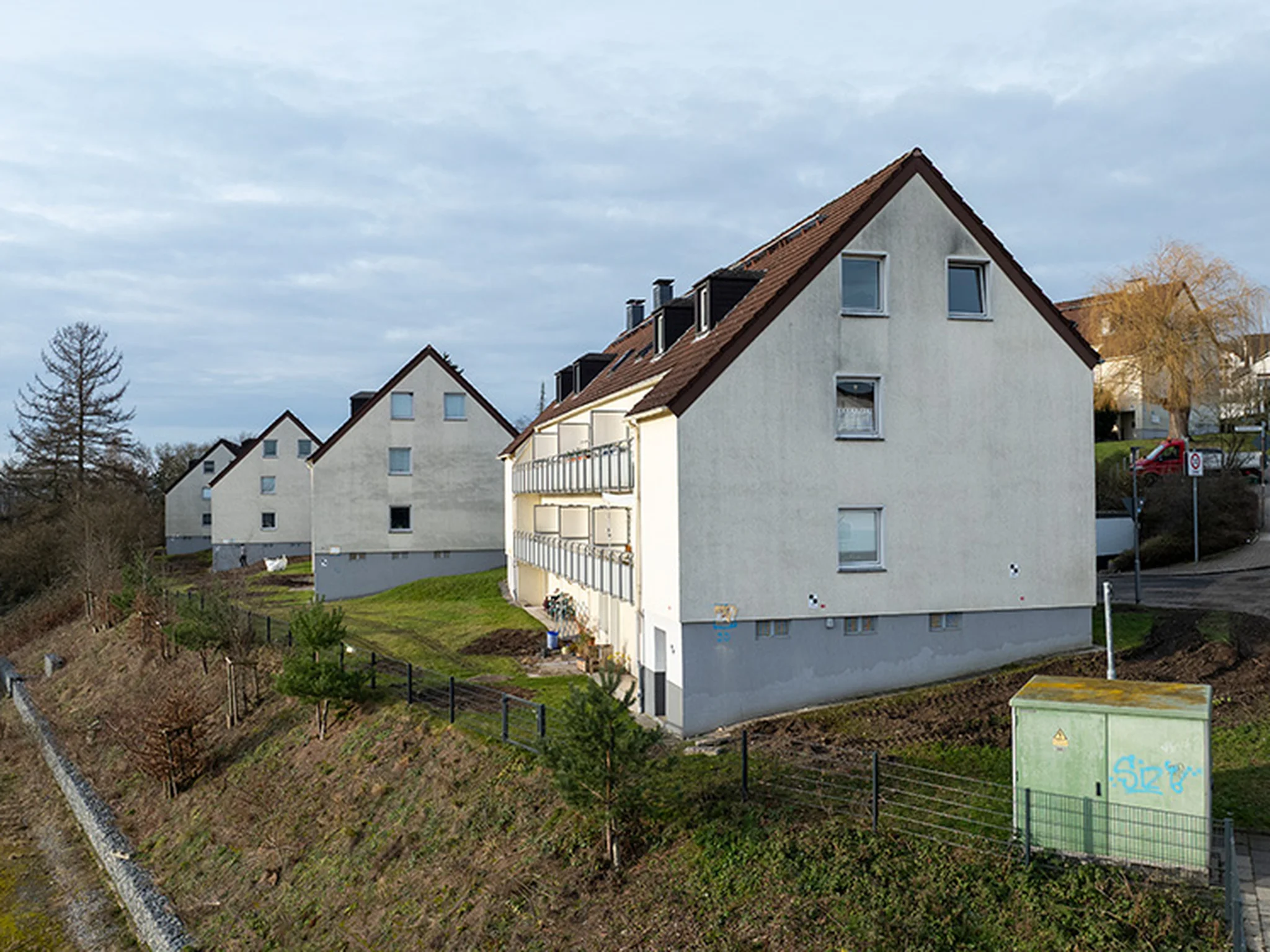 A row of three white, multi-story residential buildings with brown roofs on a sloped street, surrounded by grass and leafless trees.