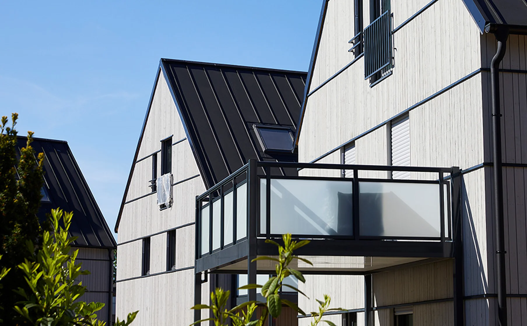 Modern light-wood houses with steep black metal roofs and frosted-glass balcony panels under a clear blue sky.
