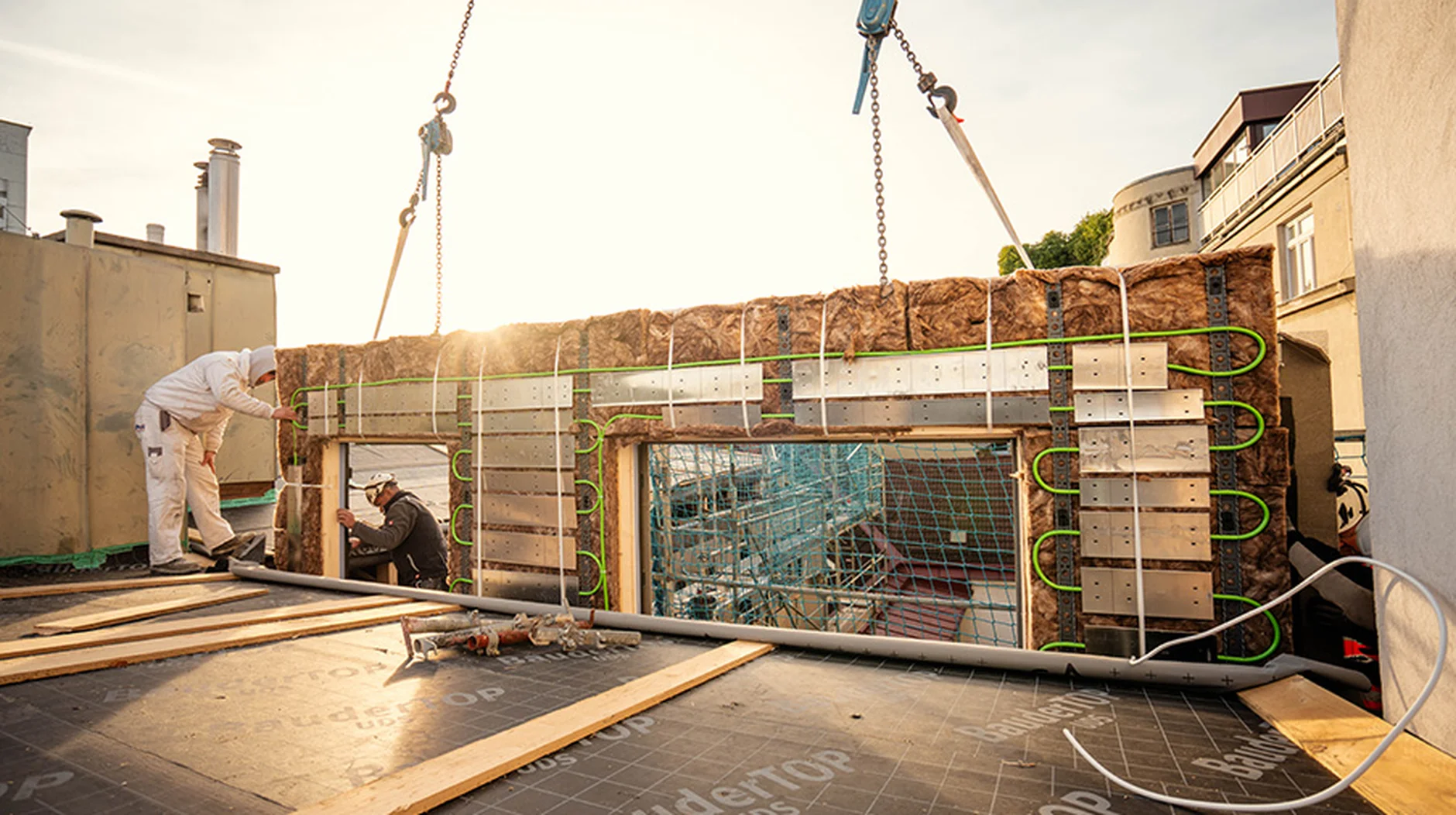 Construction workers guide a crane-lifted prefabricated insulated wall panel with visible green heating pipes into place on a rooftop.
