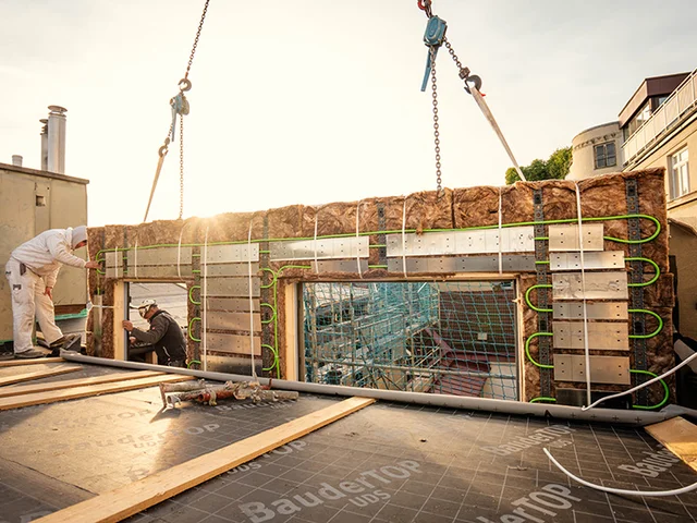 Construction workers guide a crane-lifted prefabricated insulated wall panel with visible green heating pipes into place on a rooftop.