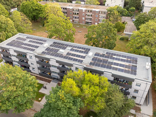 Aerial view of a multistory building with solar panels on the roof, surrounded by trees and greenery, with another building visible in the background.