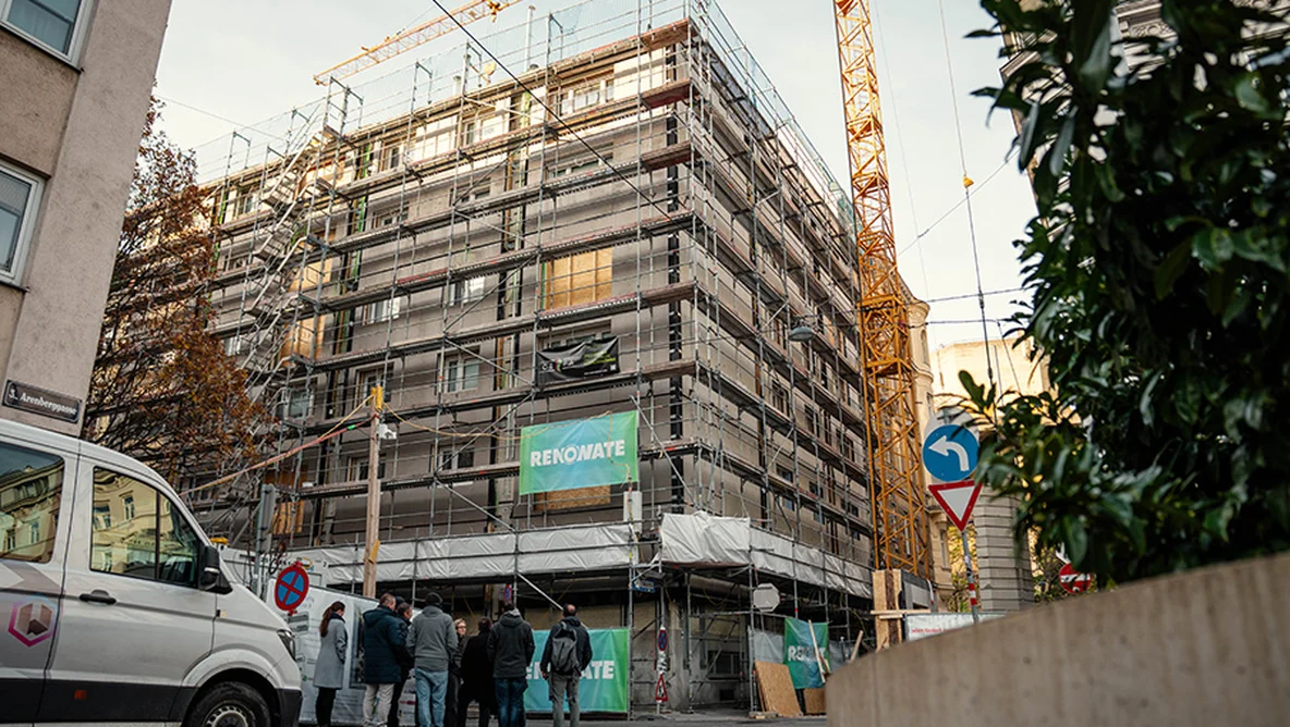 Multi-story building wrapped in scaffolding and 'RENOWATE' banners, construction crane overhead, van and group of people on the street.