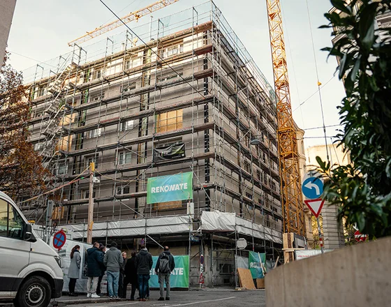 Multi-story building wrapped in scaffolding and 'RENOWATE' banners, construction crane overhead, van and group of people on the street.