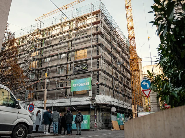 Multi-story building wrapped in scaffolding and 'RENOWATE' banners, construction crane overhead, van and group of people on the street.
